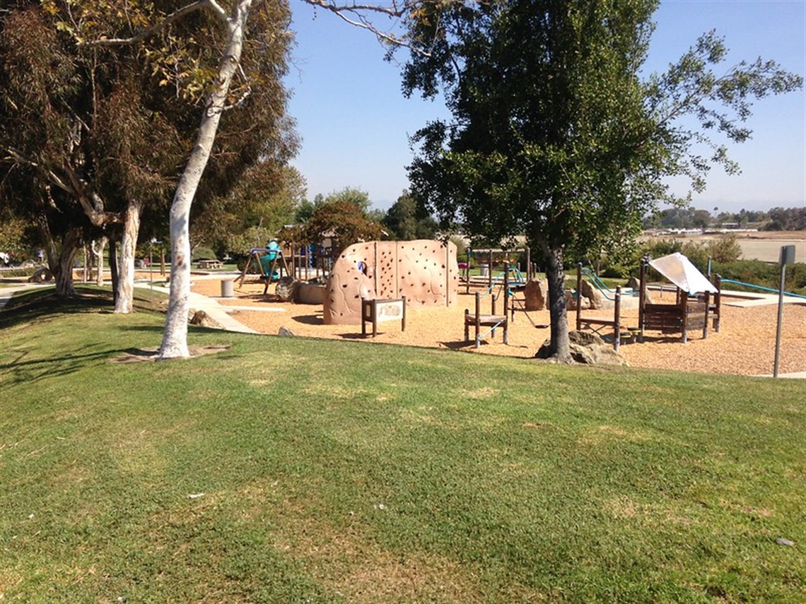 Creekside Park various play structures, including a climbing wall and slides. The playground is surrounded by trees, and the ground is covered with grass and wood chips. 