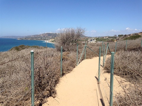 Dana Point preserve with sandy path with green poles and wire fencing, leading to the ocean.