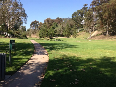 A paved pathway winds through Dana Woods Park area with trees and hills in the background. Benches and signs are along the path, with scattered leaves on the ground under a clear blue sky.