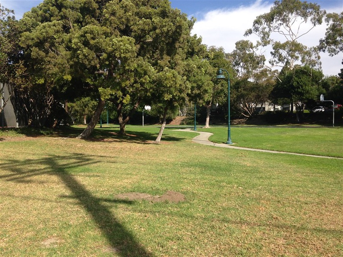Del Obispo Park with green grass, several trees, a paved pathway, and a lamp post. The shadow of an overhead structure is visible on the grass.