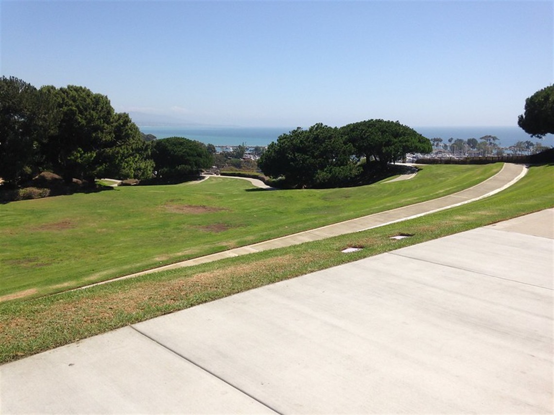 Heritage Park with green grass, trees, and a winding pathway. The ocean is visible in the background under a clear blue sky.