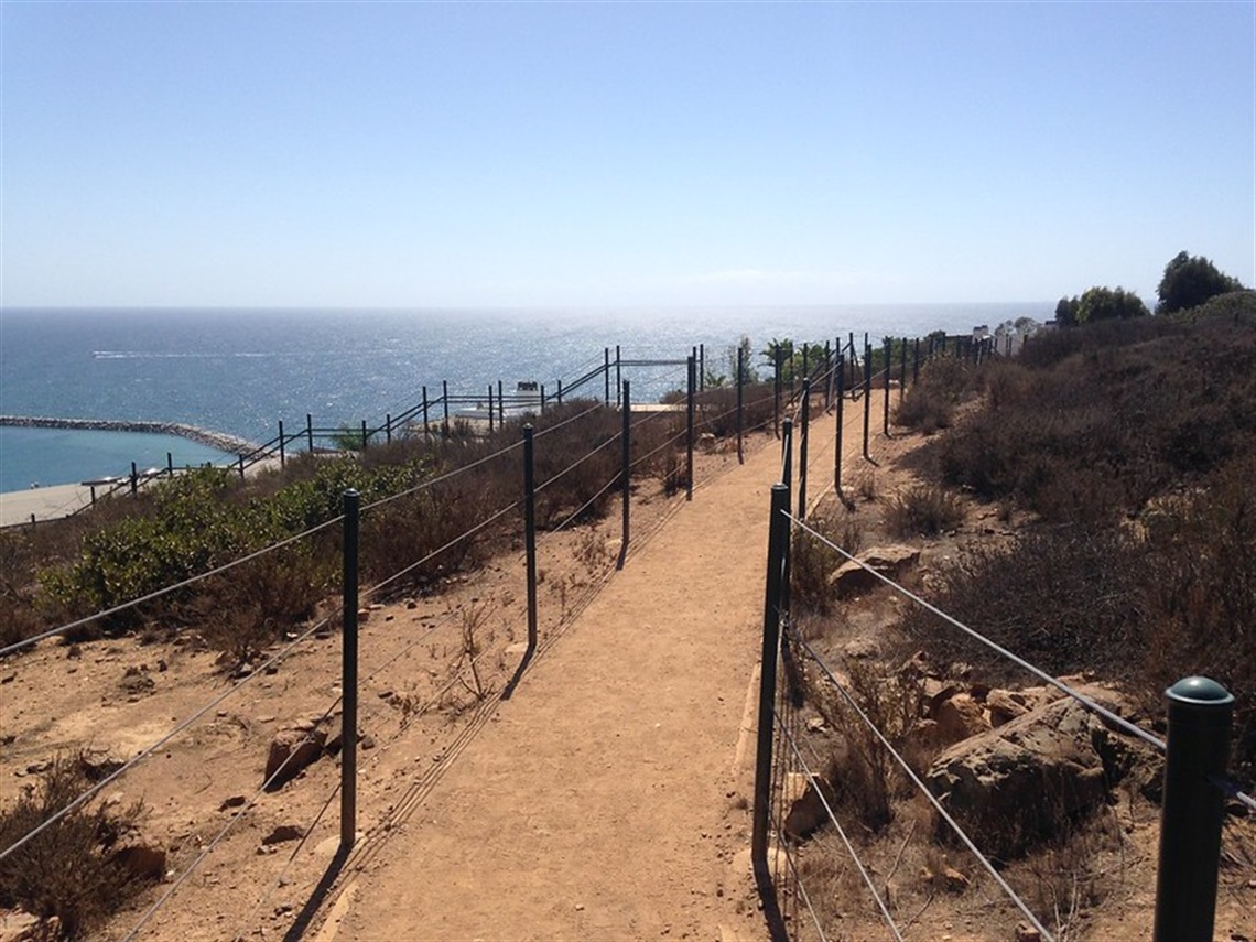 Headland Trail overlooking the ocean. The sky is clear and blue, with dry bushes and rocks along the path.