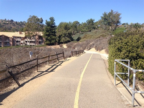 Salt Creek Bike Path showing a paved pathway with a yellow line in the center, flanked by wooden fences on the left and metal railings on the right. The path curves slightly to the right and is surrounded by dry vegetation and trees.