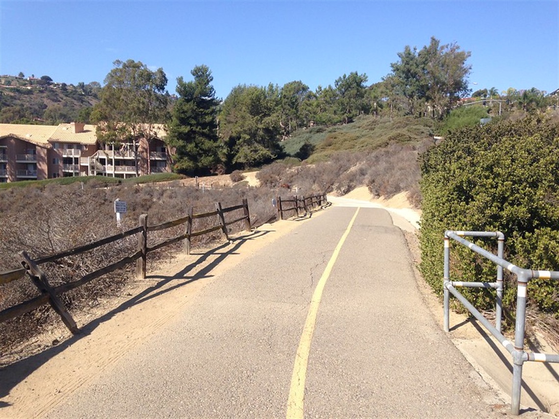 Salt Creek Bike Path showing a paved pathway with a yellow line in the center, flanked by wooden fences on the left and metal railings on the right. The path curves slightly to the right and is surrounded by dry vegetation and trees.