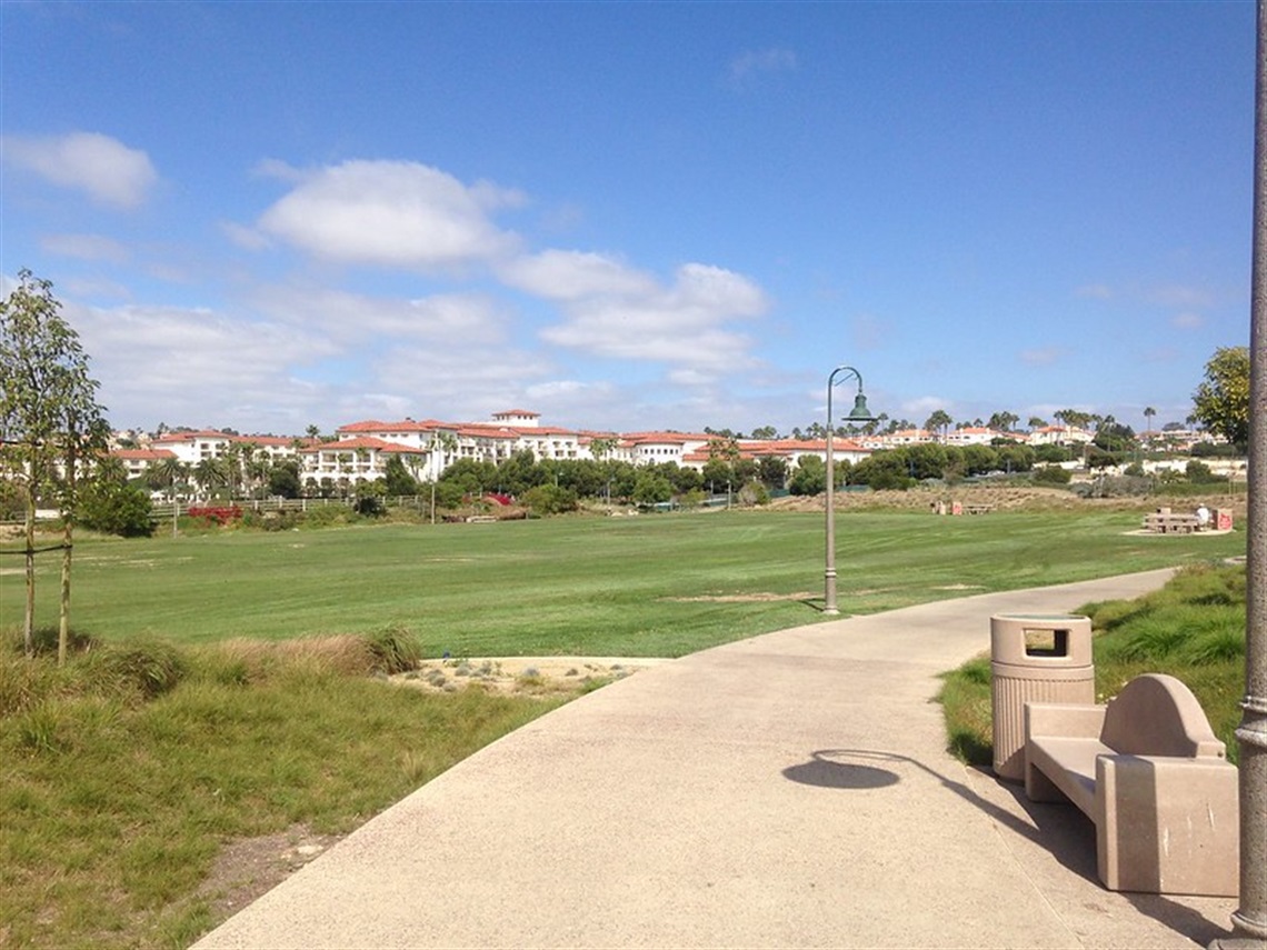 A paved pathway in Sea Terrace Park with a bench on the right side, leading towards an open grassy area with trees and lamp posts. Buildings with red-tiled roofs are visible in the background under a partly cloudy sky.