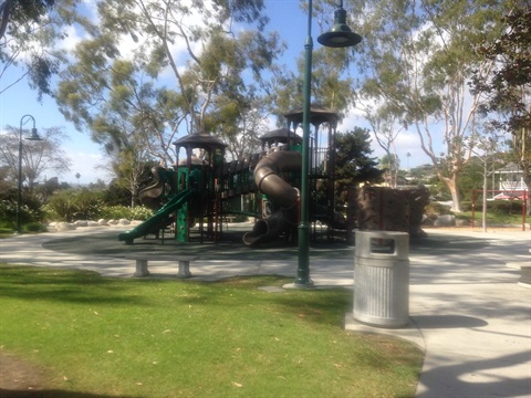 Sunset Park with various play structures, including slides and climbing equipment. There are trees in the background, a trash can in the foreground, and a lamp post near the play area.