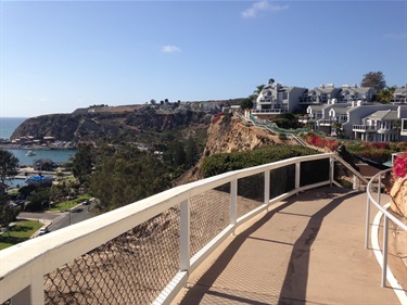 Bluff top trail coastal view from a curved walkway overlooking cliffside homes, lush greenery, and Dana Point harbor below.