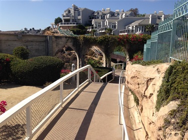 Bluff Top Trail's sloped walkway with white railing leading toward an archway covered in greenery, with houses on a hill in the background.
