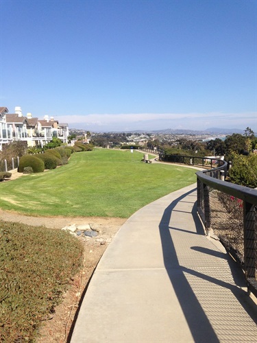 Curved concrete pathway with a metal railing overlooking a large grassy area and residential buildings, with distant hills under a clear blue sky.