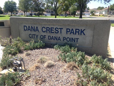 Concrete sign reading ‘Dana Crest Park – City of Dana Point,’ surrounded by gravel and small plants, with trees and houses in the background.