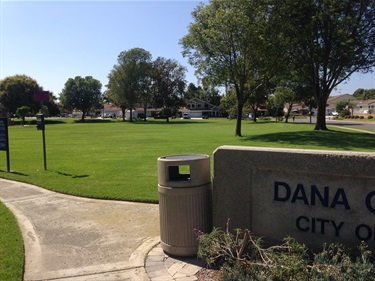 “Concrete park sign partially visible reading ‘Dana Crest Park – City of Dana Point,’ next to a trash can and curved pathway through a grassy area with trees.