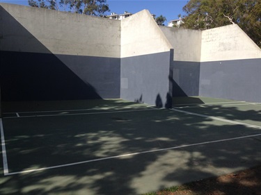 Outdoor handball court at Del Obispo Park with tall concrete walls painted in light and dark gray, and a green playing surface partially shaded by trees.