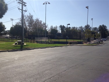 Street view of Del Obispo Park showing a baseball field enclosed by a chain-link fence, with light poles, trees, and benches along the sidewalk under a clear sky.