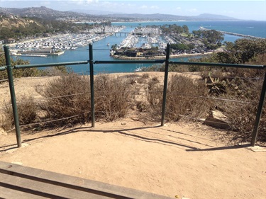 Dirt overlook with a metal railing facing Dana Point Harbo, surrounded by hills and buildings under a partly cloudy sky.