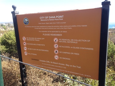 Brown sign reading “City of Dana Point – Welcome to Harbor Point Park” with park rules listed below. Surrounded by greenery, with ocean visible in the background under clear skies.