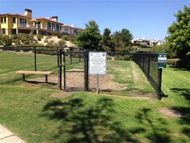 Small fenced dog park area with a bench and posted rules sign, surrounded by grass and landscaping, with residential buildings on a hillside in the background under a clear blue sky.