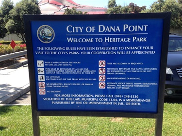 Blue sign for City of Dana Point at Heritage Park listing park rules and contact information, placed on grass with trees and a building in the background.