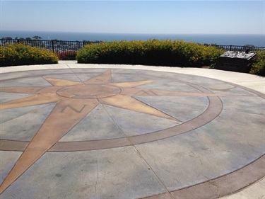 Compass rose on a circular paved overlook with ocean view and shrubs in the background.