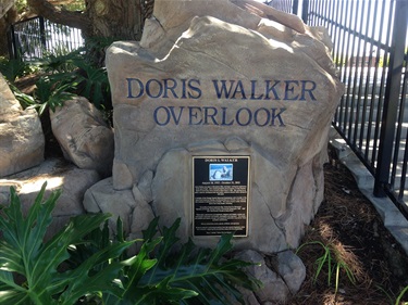 Stone monument engraved with the words “Doris Walker Overlook,” featuring a plaque with text and an image, surrounded by greenery and a black metal fence in the background.