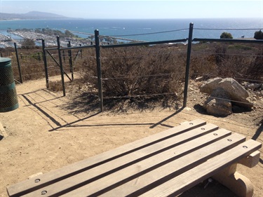 Wooden bench on a dirt overlook enclosed by railing. The viewpoint faces the ocean and Dana Point Harbor in the distance, with dry shrubs and rocks in the foreground under clear skies.