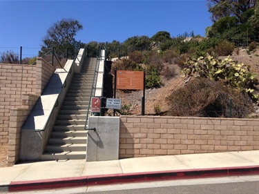 Concrete stairway with green railings ascending a hillside, bordered by a tan brick retaining wall and desert vegetation, with informational signs at the base under a clear blue sky.