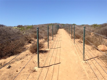 Straight dirt trail bordered by railing, cutting through dry shrubs. The path leads uphill toward a clear blue sky with no visible structures in the distance.