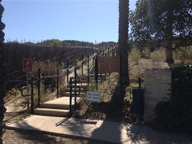 Outdoor stairway with railings leading uphill through a natural area, flanked by signs and vegetation, under a clear blue sky.