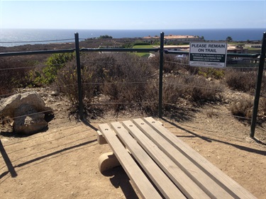 Wooden bench on a dirt surface near a wire-fenced overlook, with a sign reading “Please Remain on Trail” and ocean views in the background.