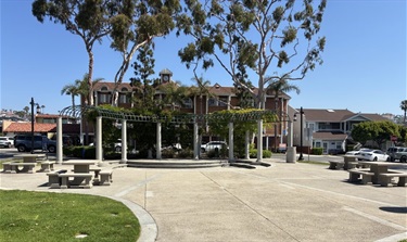 La Plaza Park gazebo-style pavilion, picnic tables, and trees surrounded by sidewalk paths and nearby buildings.