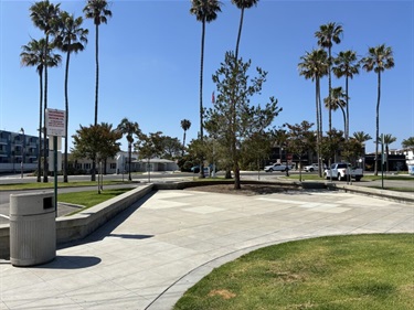 La Plaza Park curved concrete plaza with low seating walls, trees, and a trash can, surrounded by grass, palm trees, and nearby buildings and parked cars.