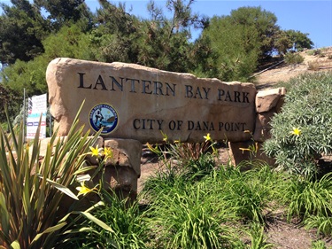 Stone sign engraved with the words “Lantern Bay Park – City of Dana Point,” surrounded by green plants and yellow flowers, with trees and shrubs in the background under bright sunlight.