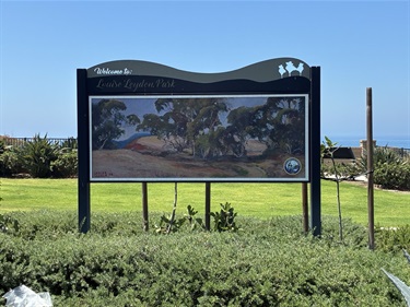 Louise Leyden Park informational signboard displaying a park rules panel and illustrative images, standing on posts in a grassy area with low shrubs and open sky in the background.