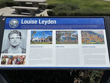 Interpretive sign titled “Louise Leyden,” featuring a portrait on the left and several photos and text panels about community history and contributions, mounted outdoors with picnic tables visible behind it.