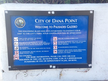 Blue sign with white text mounted on a white wall, showing park rules for Palisades Gazebo in the City of Dana Point, including welcome message, icons, and no smoking notice.