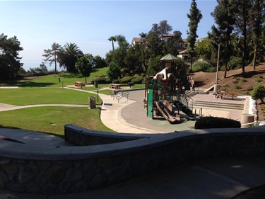 Playground area with slides and climbing structures surrounded by a paved walkway, set in a grassy park with trees and benches, under a clear blue sky.
