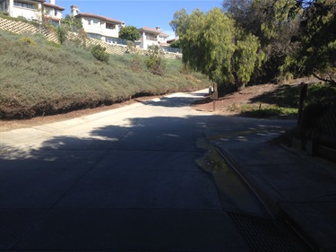 Shaded paved road curving uphill between sloped shrub-covered embankments, with houses visible along the top under a clear blue sky.