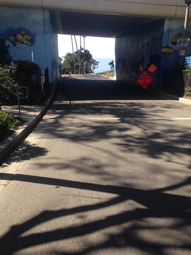Coastal view of a sandy beach with gentle waves, a paved path in the foreground, and hills with houses in the distance under a clear blue sky.