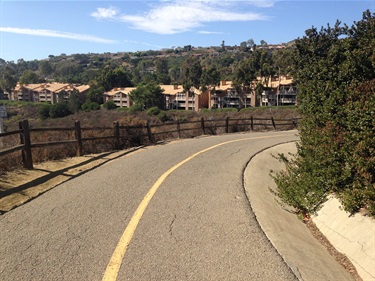 Curving paved path with a yellow center line, bordered by a wooden fence and shrubs, with hillside homes in the background under a partly cloudy sky.