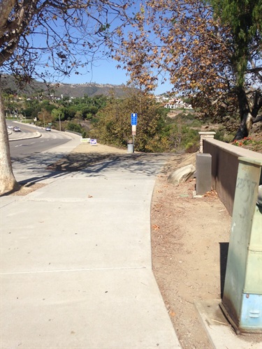 Concrete sidewalk curving alongside a road with trees and shrubs, a blue emergency call box ahead, and hills with houses in the background under a clear sky.