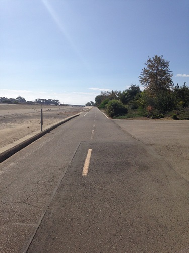 Paved trail with a yellow center line, bordered by a chain-link fence and trees on the left and a sandy channel on the right under a partly cloudy sky.