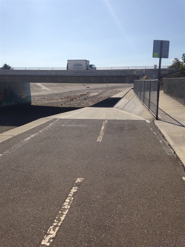 Paved trail leading under a concrete overpass with a truck visible on the road above. Chain-link fence and signpost on the right, open sandy area on the left under a clear sky.