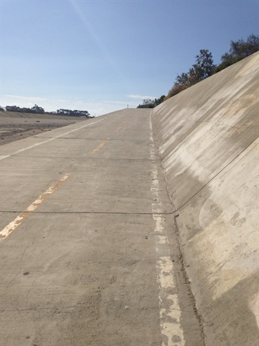 Paved incline with a yellow center line running alongside a tall concrete wall, trees visible at the top under a clear sky.