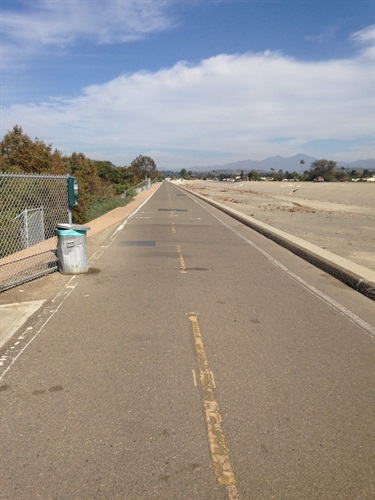 Straight paved trail with a yellow center line, bordered by a chain-link fence and trash bin on the left, and a sandy channel on the right under a partly cloudy sky.