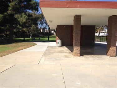 Covered picnic area with a flat roof supported by stone columns, next to a concrete walkway and grassy lawn with trees in the background.