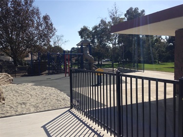Black metal fence in front of a sandy play area, with playground equipment and trees visible in the background under bright sunlight.