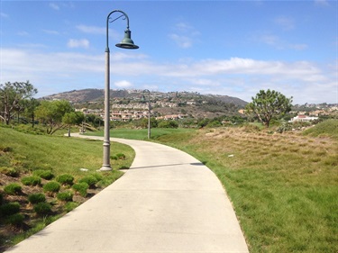 Concrete pathway winding through a grassy park with a lamp post, hills, and houses visible in the background under a partly cloudy sky.
