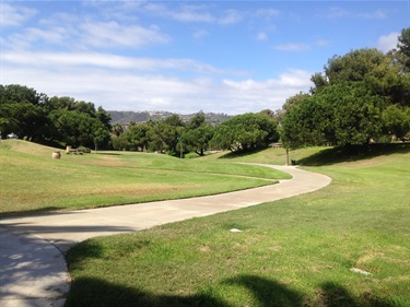 Concrete pathway curving through a wide grassy park with scattered trees and hills in the background under a partly cloudy sky.