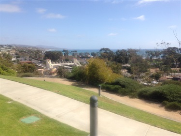 Concrete pathway in a grassy park overlooking trees, buildings, and the ocean in the distance under a partly cloudy sky.