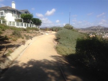 Dirt pathway curving uphill with shrubs and greenery on both sides, a large house with a white fence on the left, and hills visible in the background under a partly cloudy sky.