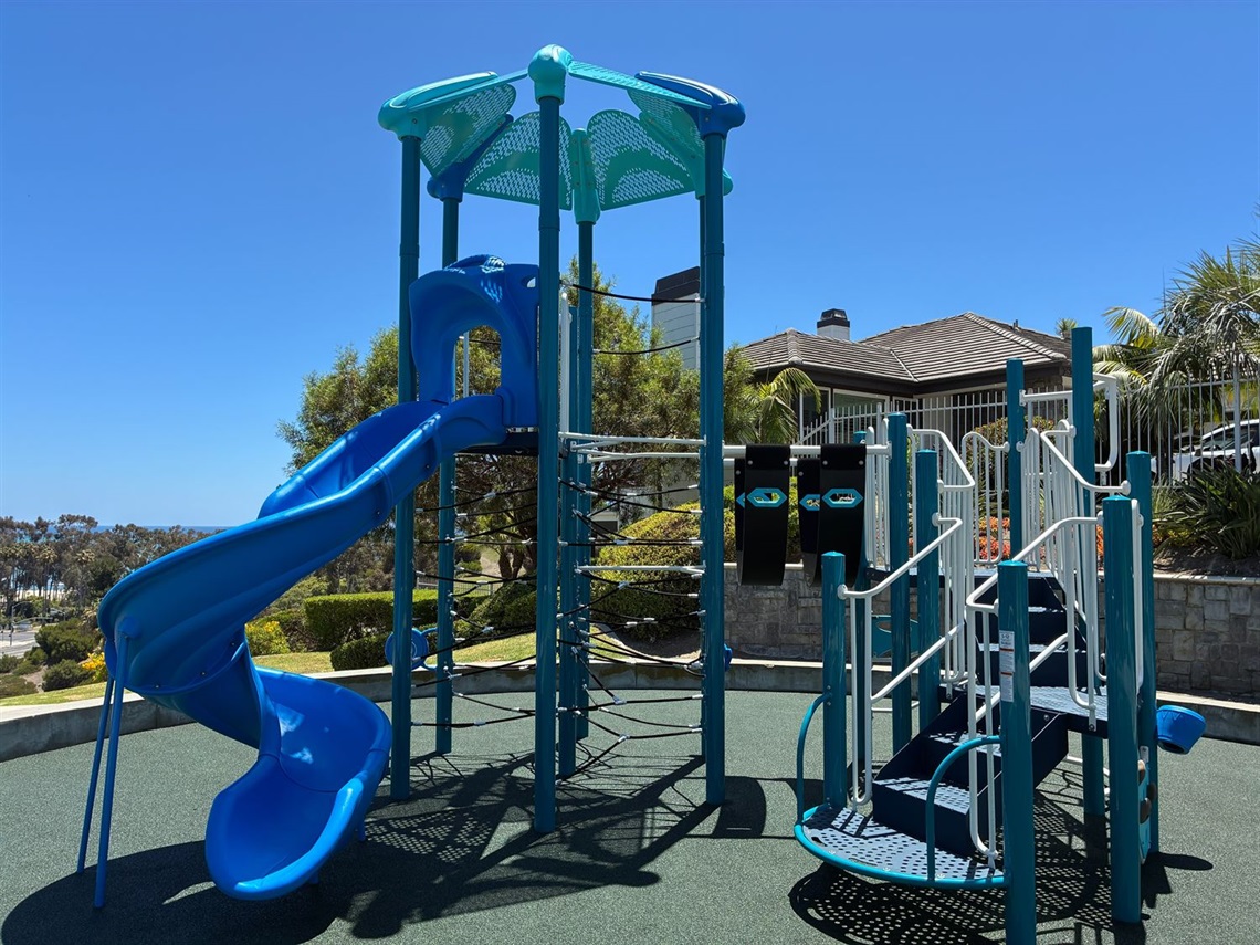 Blue playground structure with a spiral slide, climbing platforms, and stairs in an outdoor park on a sunny day.